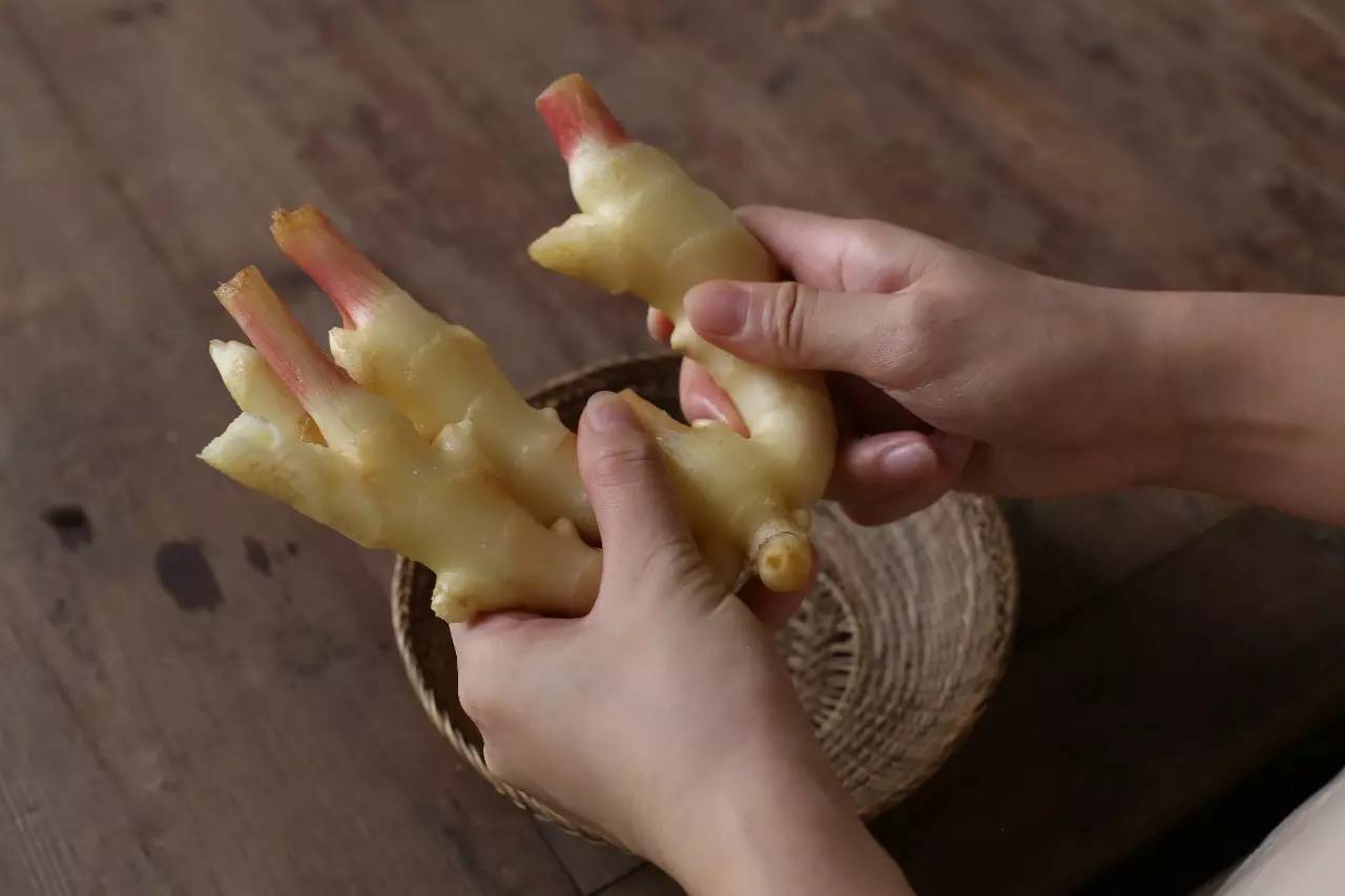 一位中老年妇女站在厨房操作台前,正在用手掰开一块带有绿色嫩芽的老姜,旁边放着菜刀和葱段,背景是整洁的厨房橱柜,阳光透过窗户洒入,营造出温馨的生活氛围 厨房场景中老人用手掰开发芽老姜