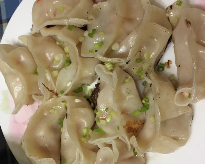 成品小笼包与饺子对比摆盘：左侧3只晶莹剔透的小笼包（皮薄透光，隐约见粉嫩肉馅，顶部点缀翠绿小葱末），右侧3只琥珀色煎饺（底部焦脆金黄，葱香油润，散发热气），背景为原木砧板