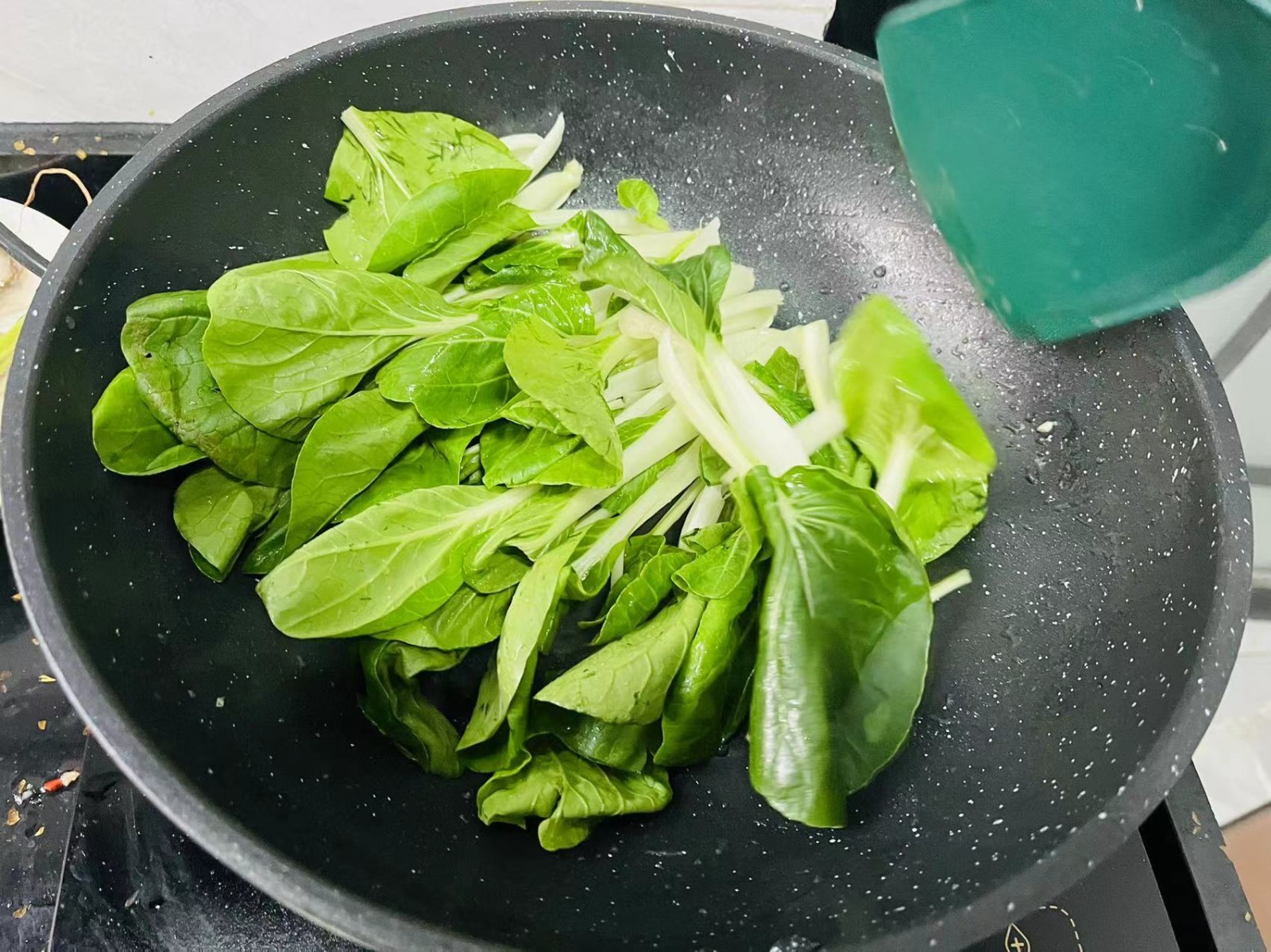 一位家庭主妇正在厨房灶台前翻炒锅中的红薯尖,锅中蔬菜呈鲜绿色,配有蒜片和红椒点缀,厨房环境整洁,窗外透进自然光,体现健康家常饮食氛围 家庭厨房炒菜场景