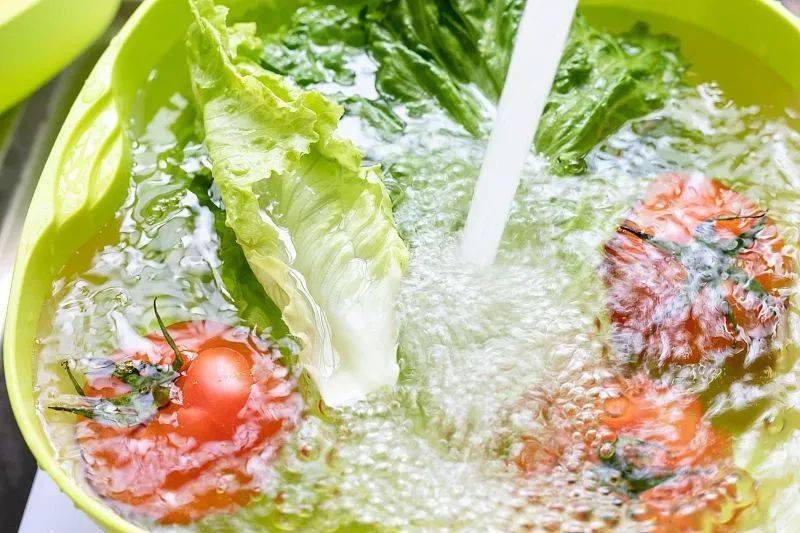 一位厨师正在用流水冲洗一把散开的深绿色散叶生菜，水珠溅起，叶片清晰可见，旁边放置着一个透明盆子盛放已洗净的生菜