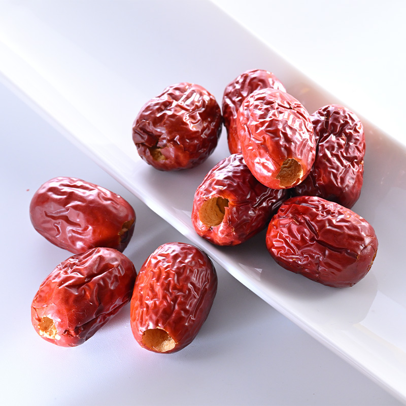 一颗完整的省枣果实特写,展示其深红色泽、光滑表皮与长椭圆形外观;背景为自然光照下的果园环境,果实置于木质托盘上,旁边配有比例尺 骏枣“省枣”特写