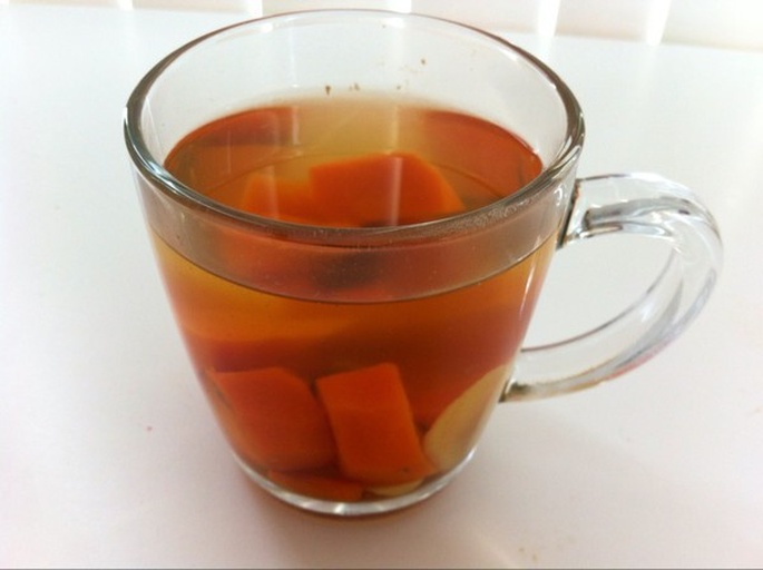 一杯透明玻璃杯中盛有淡粉色的红皮萝卜蜂蜜饮品,液体清澈微带果肉沉淀,杯口贴有一片红皮萝卜薄片作为装饰,背景为木质餐桌和蜂蜜罐 红皮萝卜蜂蜜饮成品展示