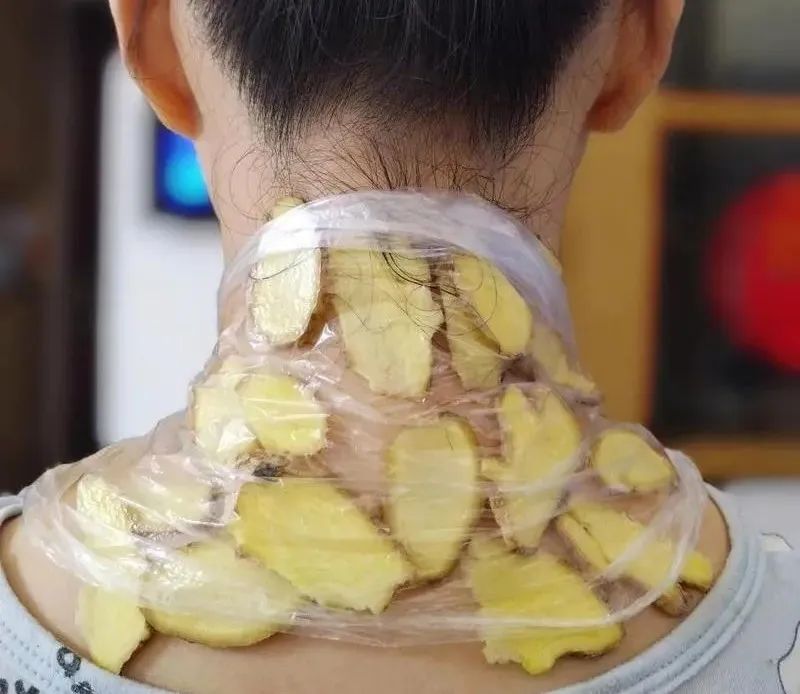 生活场景图:一位中年女性坐在木质椅子上,左手轻托右肘,右手正将一块经粗盐干炒至微黄、边缘微焦的老姜片贴敷于右侧肩颈交接处;桌面上摆放粗盐锅、姜块、研钵及棉布巾,光线柔和自然,突出‘外用’‘热敷’‘家庭理疗’关键词 老姜外用理疗场景