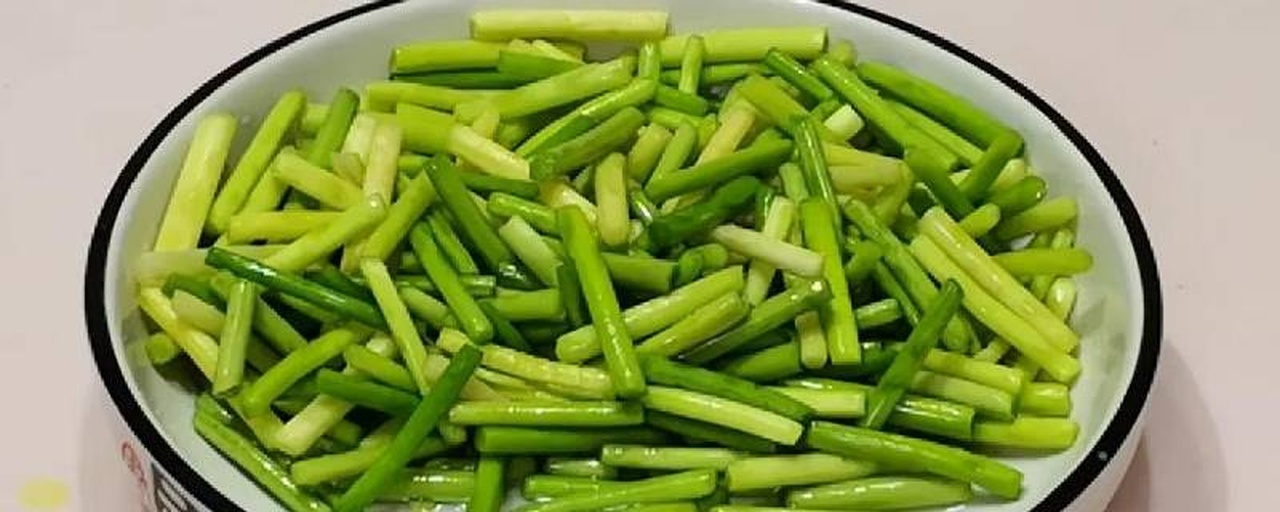 厨房场景中厨师正在切配蒜薹：不锈钢砧板上铺开洗净沥水的蒜薹段，刀工均匀约3厘米长，旁边摆放红椒丝与蒜末，光影明亮，突出食材新鲜质感与操作专业性