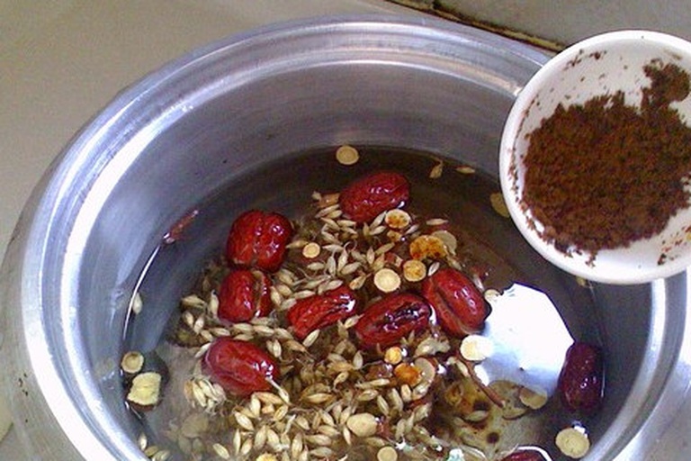 一位中年女性在厨房中用砂锅煎煮甘麦大枣汤,锅中可见漂浮的大枣、小麦颗粒和甘草片,旁边放有药材标签,标注‘淮小麦’字样,背景为木质料理台与中式橱柜,营造温馨养生氛围 煎药场景图