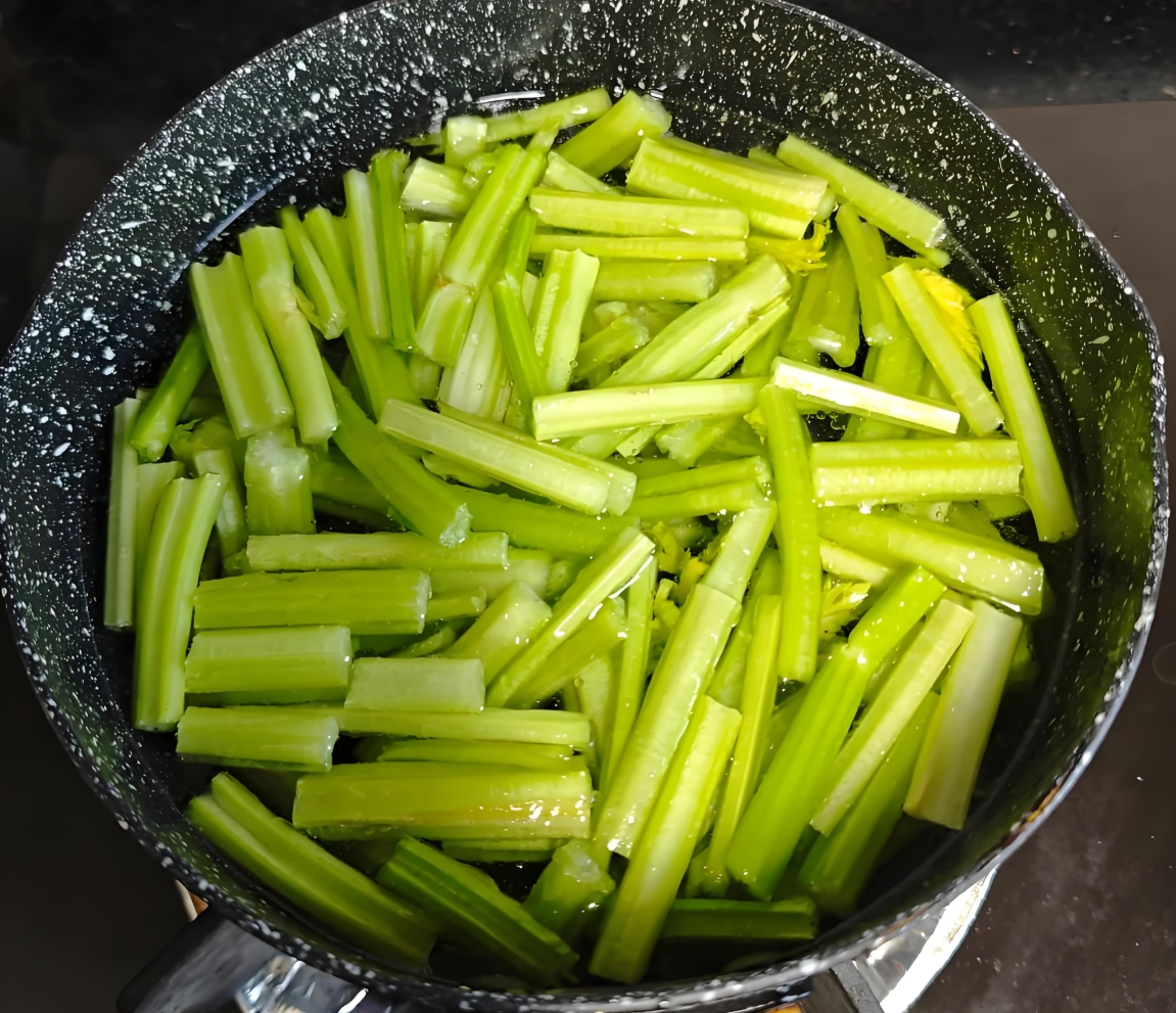 一锅清水正在沸腾,几根切成段的绿色芹菜正放入锅中焯烫,水面泛起细小气泡,芹菜颜色鲜绿,旁边摆放着漏勺和冷水盆,展示完整的焯水流程 芹菜焯水过程特写