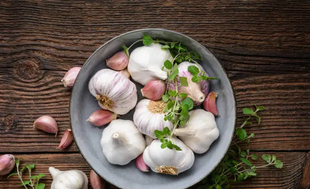木质碗中盛放新鲜大蒜,旁边配有麻布袋和透气竹篮,背景为整洁厨房橱柜,展示理想的大蒜储存环境 家庭厨房中大蒜储存场景
