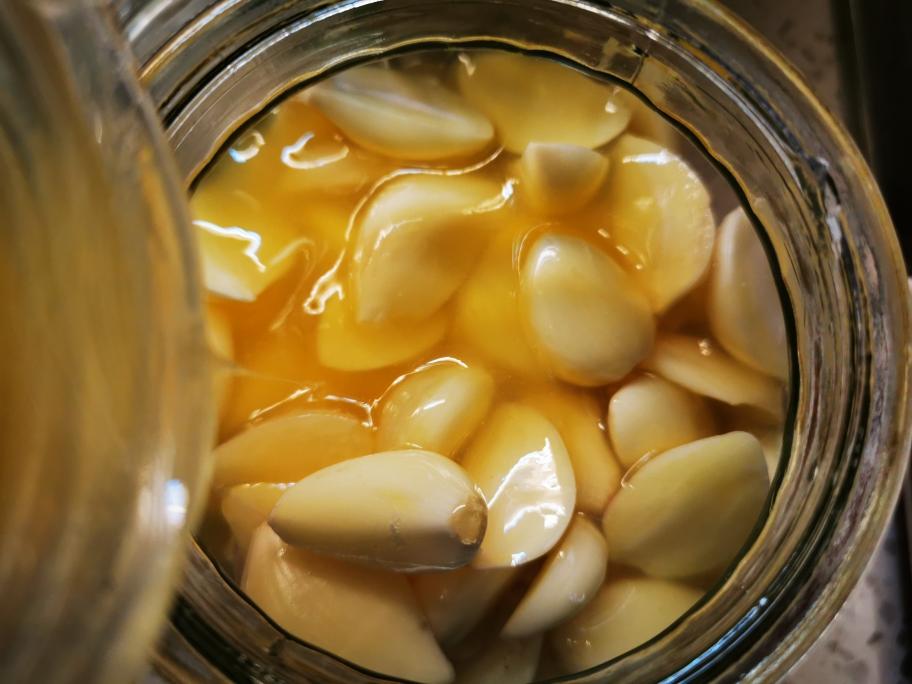 一个透明玻璃罐中盛满琥珀色的泡蒜汁,里面浸泡着洁白的蒜瓣,部分已略带微黄,罐体清晰可见气泡附着,背景为白色餐布与竹筷,整体呈现美食摄影风格 泡蒜成品展示