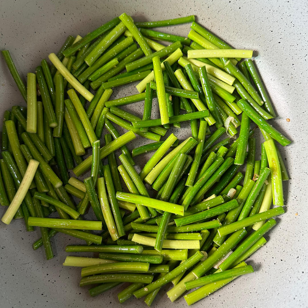 一位家庭主妇在明亮的现代厨房中用铁锅翻炒蒿子杆,锅中热气腾腾,蒜末与绿色茎叶交织,旁边放着切好的蒜片和调料瓶,展现温馨的日常烹饪画面 家庭厨房中正在翻炒蒿子杆的场景