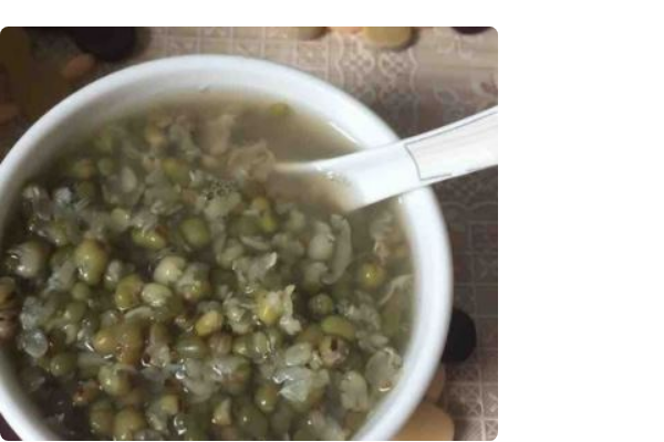 一口砂锅中翻滚着翠绿色的绿豆汤,部分绿豆已破裂露出黄色内芯,锅盖半开,热气腾腾,旁边放有一碗盛好的绿豆汤和一把木勺 一锅正在熬煮的绿色绿豆汤