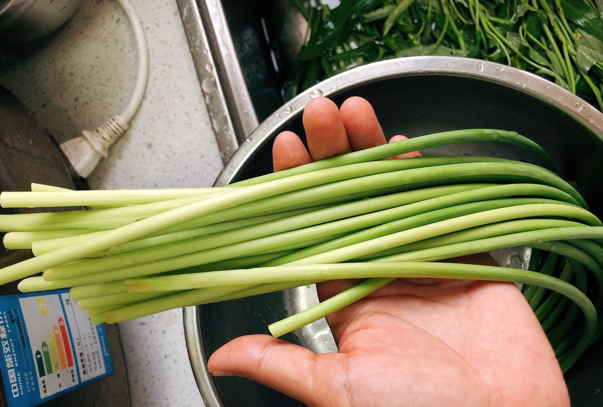 一个干净的厨房台面上,一双戴着橡胶手套的手正在用清水冲洗一把翠绿的蒜薹,旁边放着菜板和刀具,水珠溅起,背景中有调味瓶和锅具,展现家庭烹饪的安全卫生场景 厨房中正在清洗和切段蒜薹的过程