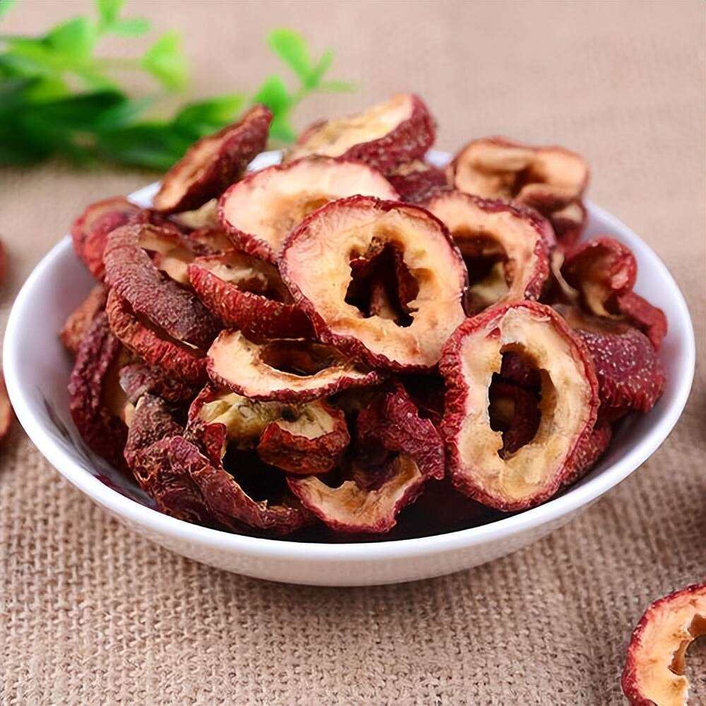 一名亚洲女性身穿居家服站在明亮的厨房中,左手端着白色瓷碗,内装约10片干红枣片,右手正将一片放入嘴中,表情自然放松,窗外晨光洒入,营造出健康生活的氛围 一位女性在清晨厨房中手持小碗食用红枣干