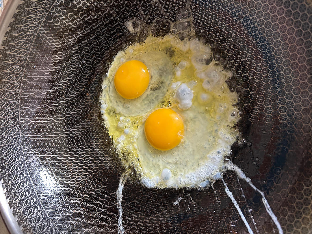 一位家庭主妇正在厨房煎鸡蛋,平底锅中金黄色的鸡蛋正在滋滋作响,旁边放着打散的鸡蛋液、盐罐和新鲜香葱,台面上还有全麦面包和一小碗糙米饭,展现鸡蛋作为蛋白质来源与主食搭配的合理膳食模式 厨房场景中的鸡蛋料理