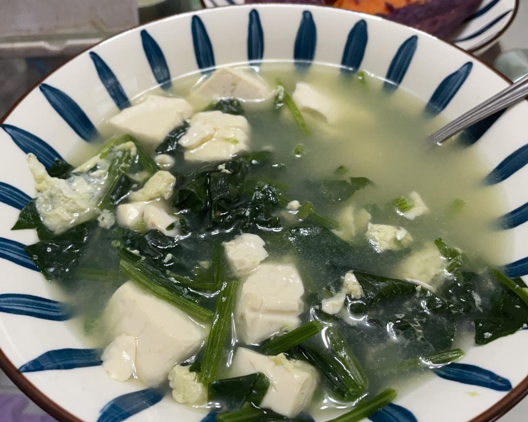 一家四口围坐在餐桌旁,每人面前摆放一碗热腾腾的菠菜豆腐汤,孩子正在用勺子喝汤,父母微笑看着,桌上还有米饭和其他小菜,氛围温暖 家庭餐桌上一家人享用菠菜汤的温馨场景