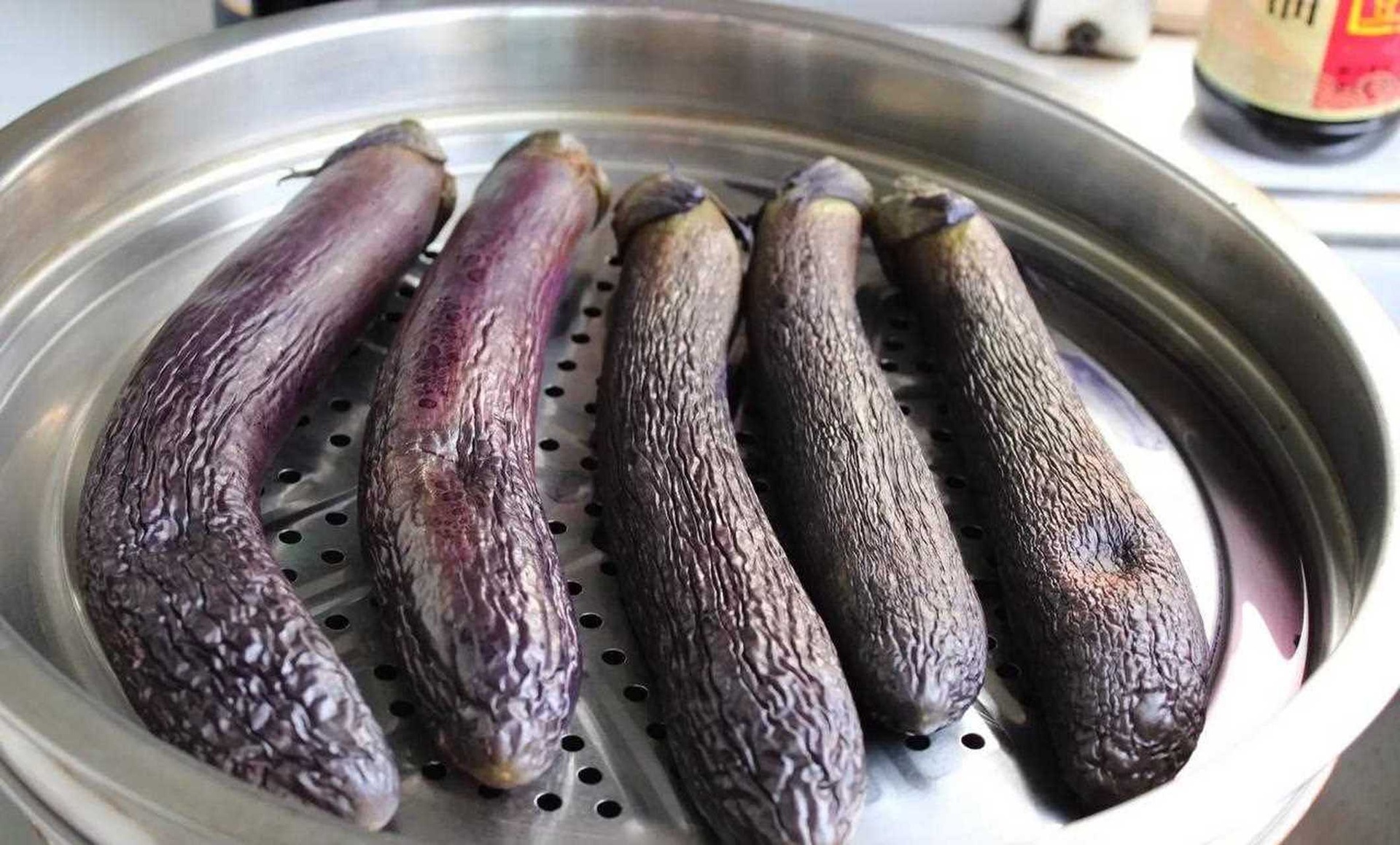 竹蒸笼蒸制长茄子实景:突出茄段单层平铺、蒸汽升腾状态及茄皮在高温下的透亮紫红光泽 蒸制过程特写:竹蒸笼中整齐平铺的茄段,蒸汽从缝隙蓬勃升腾,茄皮在高温下泛出透亮紫红色泽