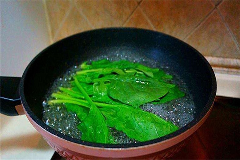 一位厨师正在厨房中将一把绿色菠菜放入冒着热气的锅中焯水,旁边配有漏勺和装有冷水的碗,展示正确的预处理方法 厨房中焯水处理菠菜的过程