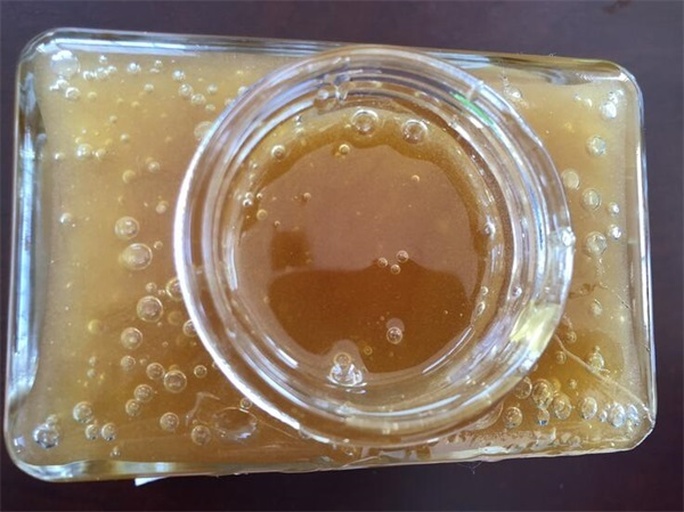 一个透明玻璃蜂蜜罐中盛满金黄色的蜂蜜,表面覆盖着一层均匀细腻的白色泡沫,光线从侧面照射,突出泡沫的质感和蜂蜜的通透性 蜂蜜瓶中漂浮细腻泡沫特写