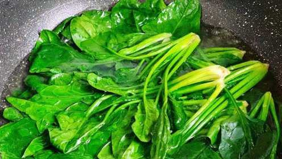 一位家庭主妇正在厨房用锅焯煮一把新鲜菠菜,锅中水沸腾,菠菜呈翠绿色,旁边放有切好的蒜末和调味料,台面整洁,光线明亮,体现健康家常烹饪氛围 家庭烹饪菠菜场景