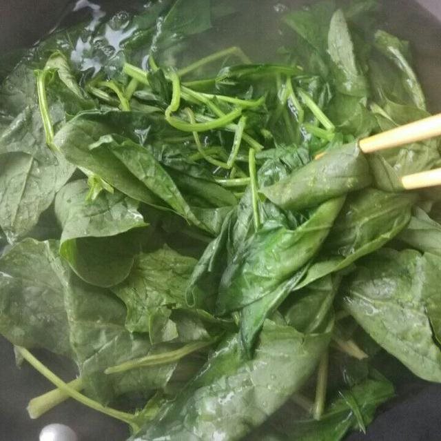 俯拍高清摄影:不锈钢锅中沸腾清水(水面有密集气泡),新鲜菠菜整株浸没,计时器显示00:60;旁边置冰水盆(水面浮冰粒),焯烫后菠菜正被捞入漏勺沥水;特写滴落水珠中可见细微白色絮状物(溶解的草酸钙微粒),印证焯水去苦有效性 家庭厨房中菠菜焯水操作实景图