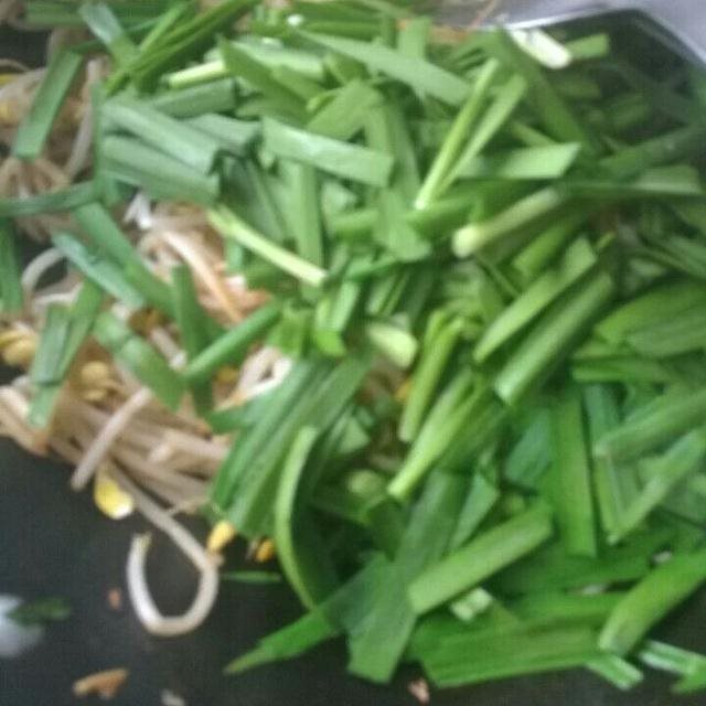 新鲜韭菜与一碗自制豆浆并列摆放，背景为厨房操作台，左侧是洗净的绿叶韭菜，右侧是一杯乳白色豆浆，旁边放有勺子和滤网