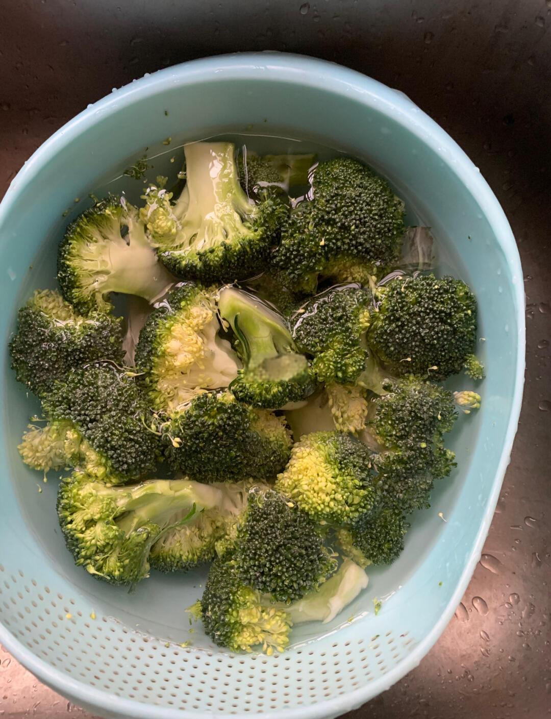 新鲜西兰花被剪成小朵,放入透明玻璃盆中的淡盐水中浸泡,背景为厨房操作台,光线自然明亮 西兰花清洗与分切过程