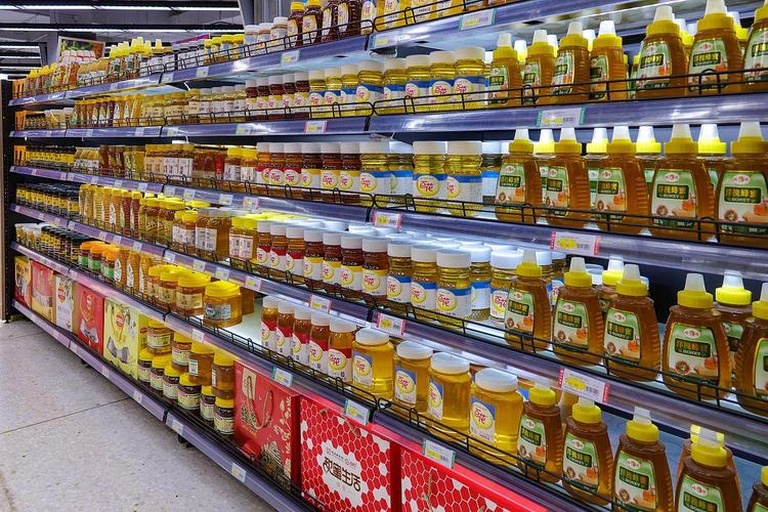 现代超市蜂蜜货架场景,多个玻璃瓶装蜂蜜整齐排列,一位女性消费者正仔细阅读某款蜂蜜瓶身上的标签,重点聚焦于营养成分表和执行标准 超市货架上摆放多种品牌蜂蜜产品,消费者正在查看标签信息