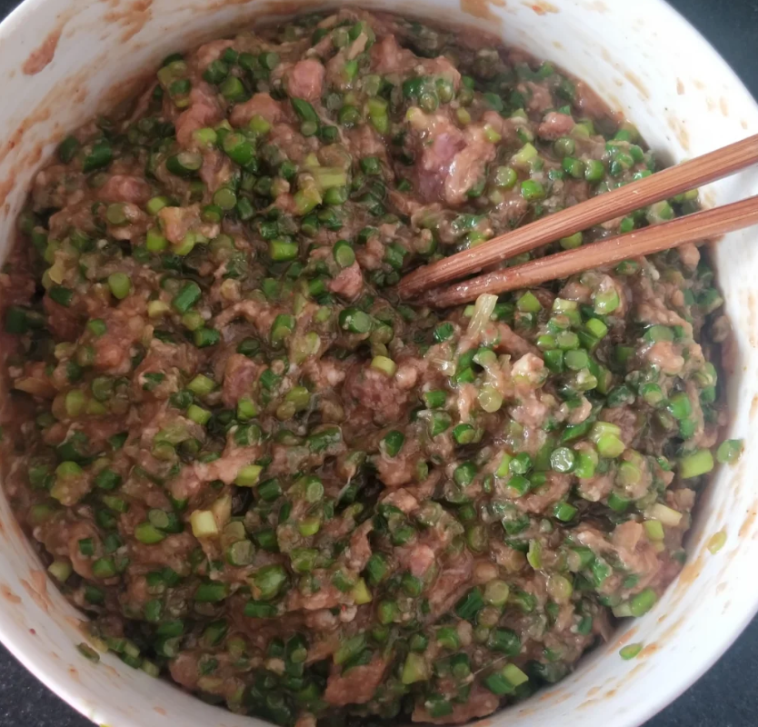 白色瓷碗中盛放着翠绿色的韭菜碎与粉红色的虾仁丁混合而成的饺子馅,虾仁分布均匀,韭菜光泽明亮,一双木筷正在轻柔翻拌 韭菜与虾仁混合馅料