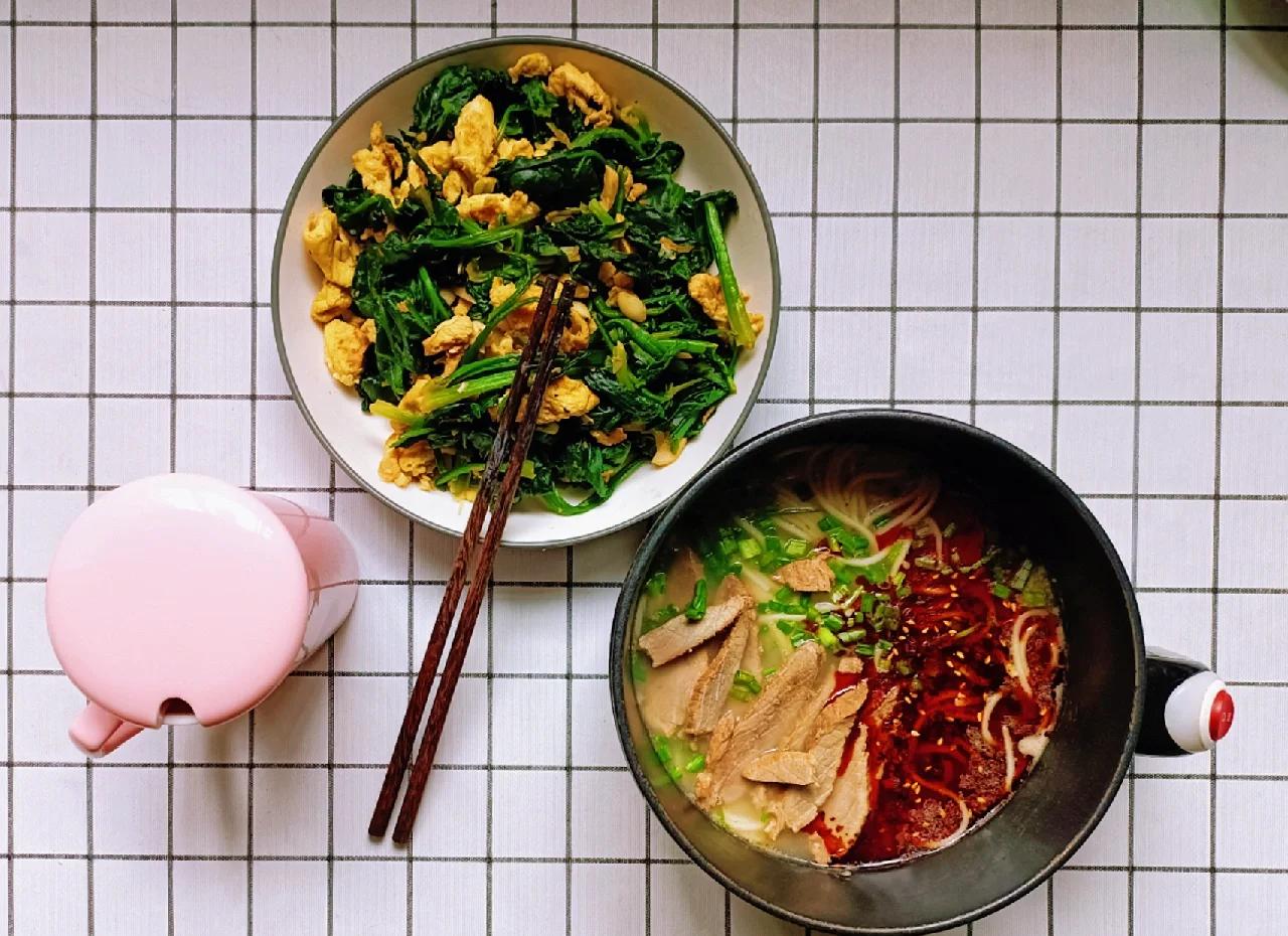 一碗热气腾腾的菠菜鸡蛋面特写，面条上覆盖翠绿菠菜与金黄鸡蛋块，汤汁清澈，配有勺子与筷子，背景虚化突出主体