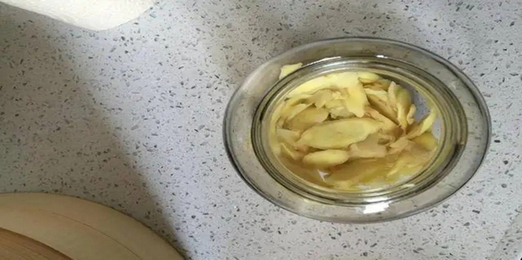 玻璃罐中盛放着浅粉色的腌渍子姜,液体清澈微泛光泽,周围散落几片新鲜子姜和一瓶米醋,背景为白色瓷砖厨房台面 家庭自制泡姜过程