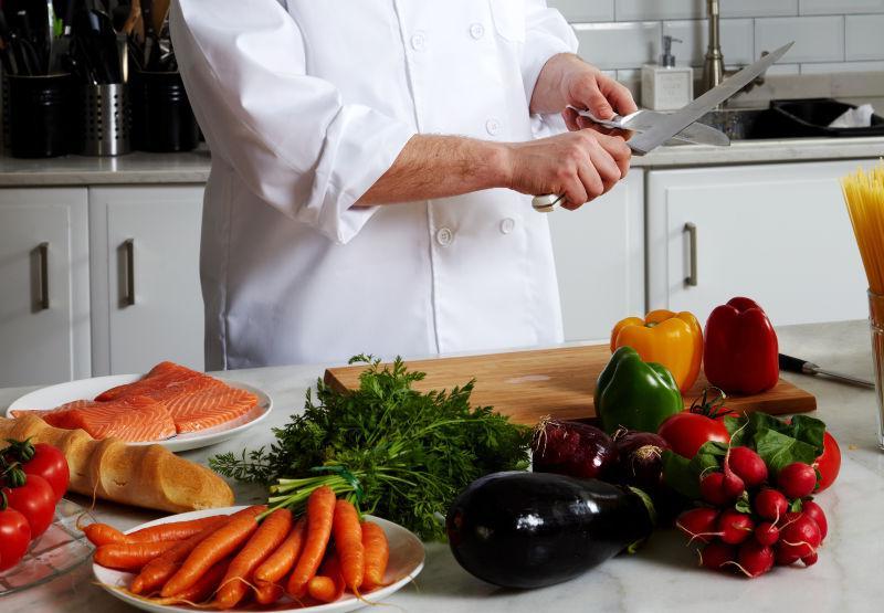 一位身穿白色厨师服的厨师在明亮的厨房中手持刀具,正在清理一条新鲜的多宝鱼,案板上有柠檬片和香草,鱼身银白带褐斑,旁边摆放着调味料瓶,背景为不锈钢厨具和炉灶 厨师正在厨房处理新鲜多宝鱼准备烹饪