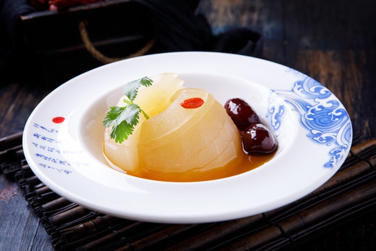 一位中年女性正在厨房用玻璃量勺称量红色宁夏枸杞，准备投入已切块的雪梨锅中