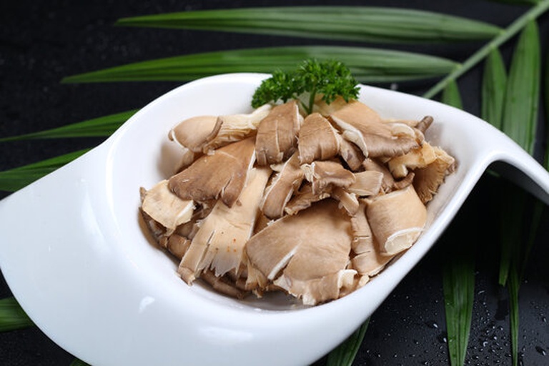 新鲜白玉菇整齐排列在白色瓷盘左侧,右侧摆放去皮切块的淡黄色土豆,旁边有剥好的大蒜、绿色青椒条和小葱段,背景为木质料理台面,光线柔和自然 食材摆放示意图