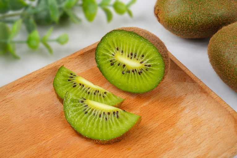 一张俯拍图片,展示绿心猕猴桃的不同食用方式:左侧为完整的带皮果实,中间为切开的两半,右侧为切成薄片摆放在白色瓷盘中;下方有一杯绿色果汁和一碗搭配蓝莓、燕麦的酸奶碗,背景简洁干净,突出健康生活方式的主题 绿心猕猴桃多种吃法展示