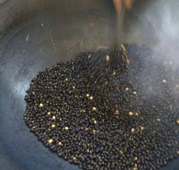 炒制黑豆过程实拍图,展示文火翻炒状态 炒锅中正在翻炒黑色豆粒的特写镜头,豆子表面微泛油光,有轻微烟气上升,背景为厨房灶台环境