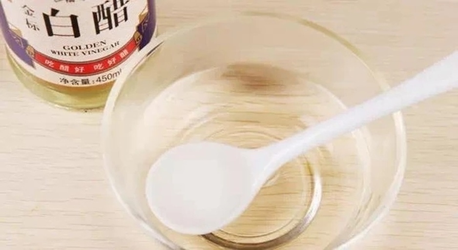 一位厨师正在玻璃碗中调制油醋汁,手持打蛋器顺时针搅拌,液体呈现乳白色乳化状态,旁边摆放着柠檬、蜂蜜罐、橄榄油瓶和研磨胡椒器,桌面铺有亚麻餐布 菠菜沙拉调味过程