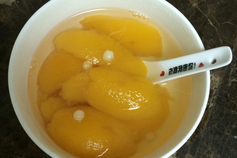 黄桃在陶瓷锅中被清水和冰糖慢炖,表面漂浮着几颗完整黄桃块,汤汁微稠,背景有枸杞和桂圆点缀,体现传统中式甜品风格 黄桃在锅中慢火炖煮,汤色呈琥珀色,周围配有冰糖、枸杞等辅料