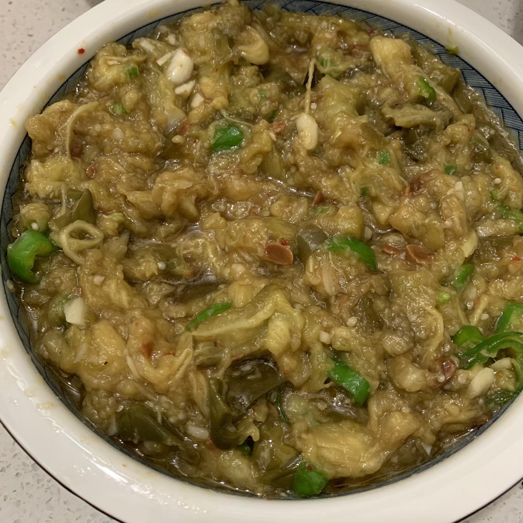 中老年女性用竹筷夹起蒜泥茄子送入口中，神情愉悦，背景为家庭厨房台面，可见紫长茄茎叶残余和带蒜皮的蒜瓣