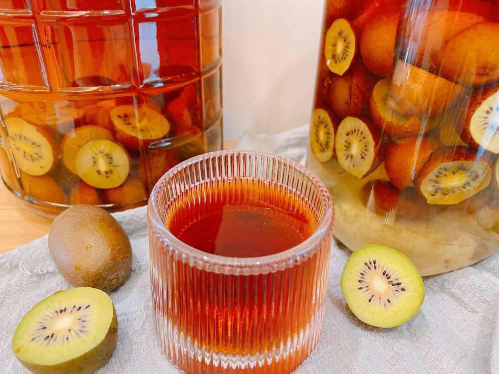 锅中正在熬煮的猕猴桃糖浆呈现出琥珀色光泽，气泡细腻，果丁悬浮其中，手持木铲轻搅，灶台为家庭厨房场景