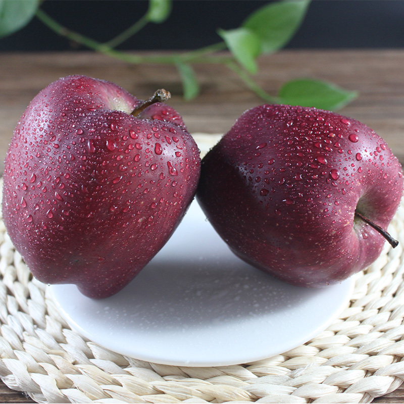 一个被横向切开的红星苹果置于白色瓷盘中,露出中心清晰的五角星状籽室结构,果肉呈乳白色,汁水丰盈。背景虚化的绿叶与红色果皮形成鲜明对比,突出其独特内部形态与新鲜质感 切开的红星苹果特写