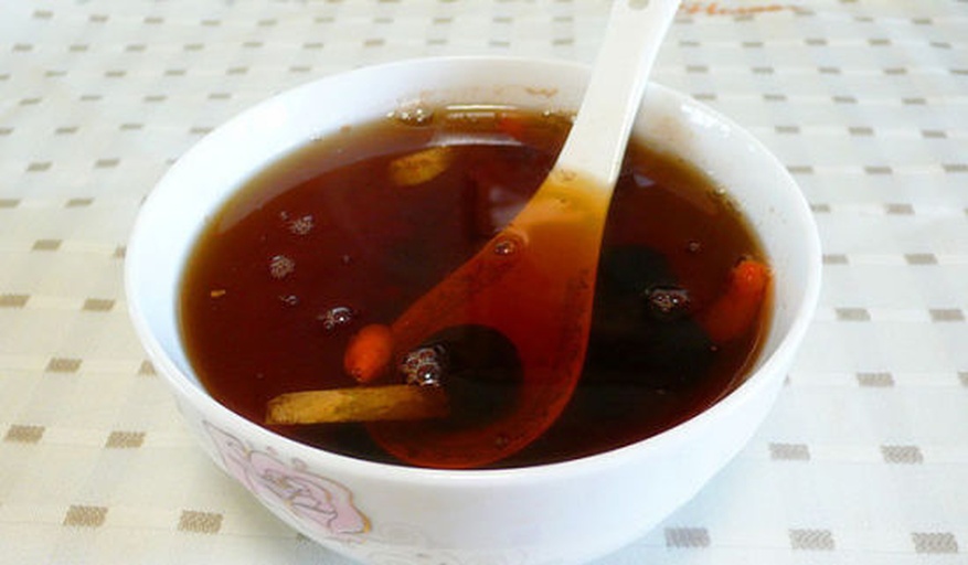 一碗热腾腾的贵妃煲被盛在白色瓷碗中,汤色呈乳白偏金黄,可见鸡肉块、红枣、枸杞与大块橙黄贵妃芒果肉,表面浮有细小油花与香草点缀,背景为木质餐桌与阳光洒落的效果,营造温馨家常氛围 成品贵妃煲盛装展示