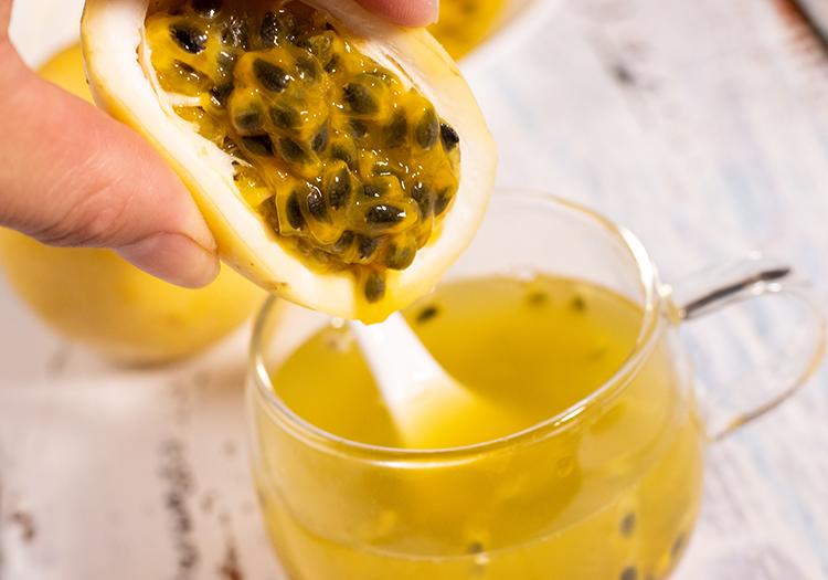 透明玻璃杯中正在倒入温水,杯底已有挖出的百香果果肉和一勺白糖,手握长柄勺轻轻搅拌,背景为厨房台面,旁边摆放切开的百香果和糖罐 百香果冲泡过程示意图