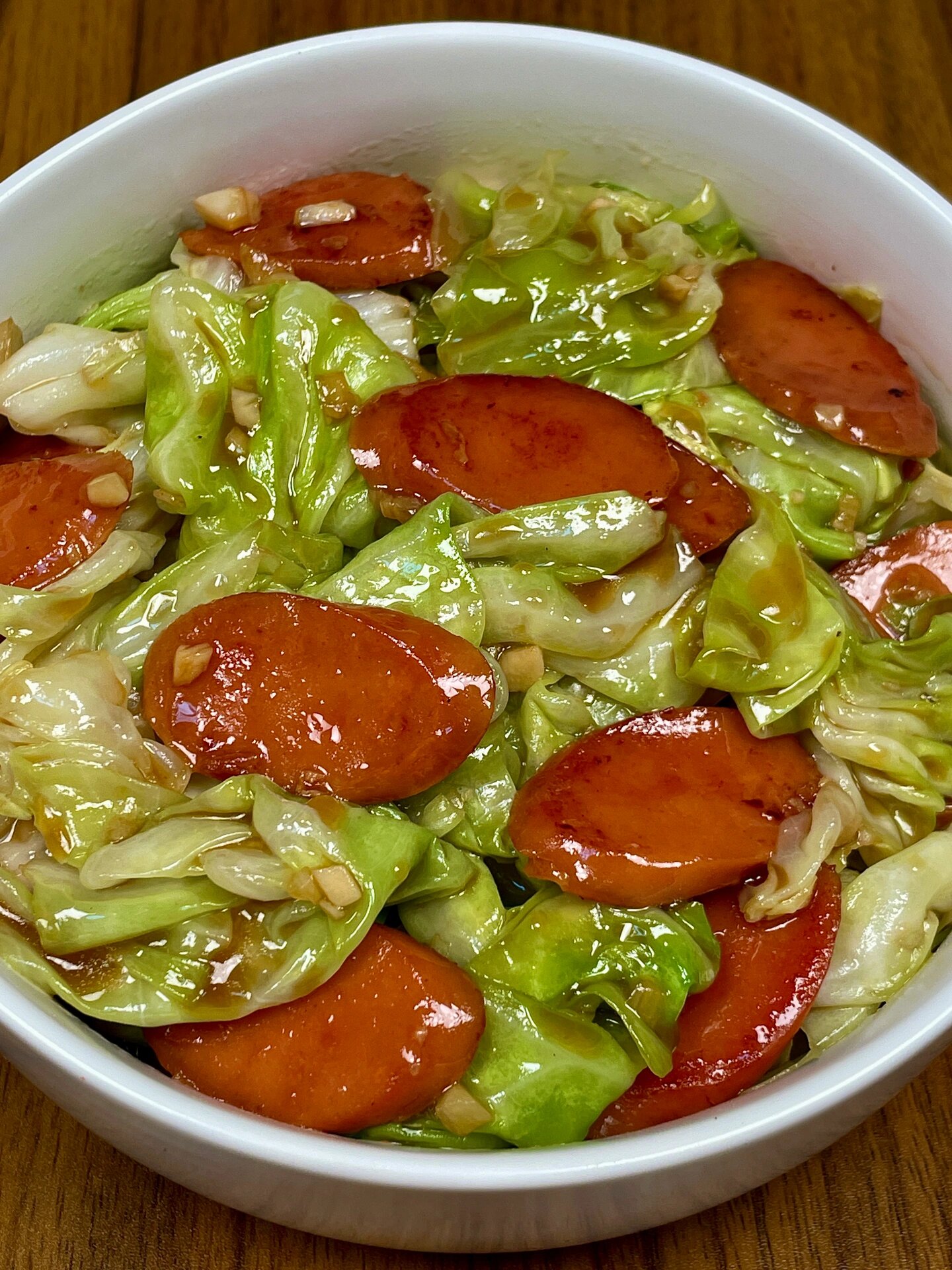一张美食特写照片,白色瓷盘中盛放着数片切开的蒜薹香肠,肉质呈玫瑰红色,内部均匀分布着翠绿色蒜薹颗粒,边缘略带油光,旁边配有胡萝卜片和香菜点缀,背景虚化,突出食物主体,光线柔和温暖,展现诱人食欲 蒜薹香肠成品菜肴图