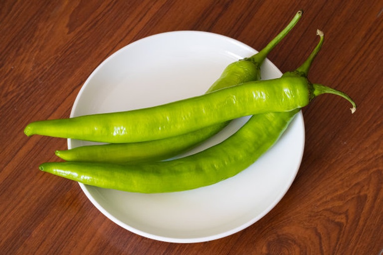 一盘新鲜采摘的青椒和彩色圆椒整齐摆放在木制桌面上,背景为自然光线下的厨房环境,青椒呈深绿色,圆椒包括红色、黄色和绿色三种颜色,表面光滑有光泽,带有少量水珠,显得非常新鲜 青椒圆椒特写