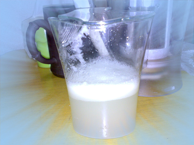 透明玻璃杯中盛有淡黄色乳状液体,表面有少量泡沫,中部可见轻微分层现象,杯壁附着细微白色絮状沉淀。杯子置于木质餐垫上,旁边放有一把金属勺子,强调饮品的真实饮用场景 一杯现榨菠萝牛奶汁呈现轻微分层状态,杯壁可见细小絮状物