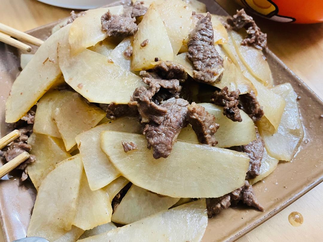 白萝卜与新鲜牛肉切片摆放在木质砧板上，旁边有葱姜蒜调料，背景为厨房灶台