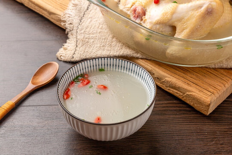 一位家庭主妇手持小勺将白胡椒粉轻轻撒入一碗冒着热气的鸡汤中,碗边配有葱花和枸杞装饰,整体画面温馨,体现食材在日常饮食中的健康应用 家庭厨房中正在将白胡椒撒入热汤中的场景