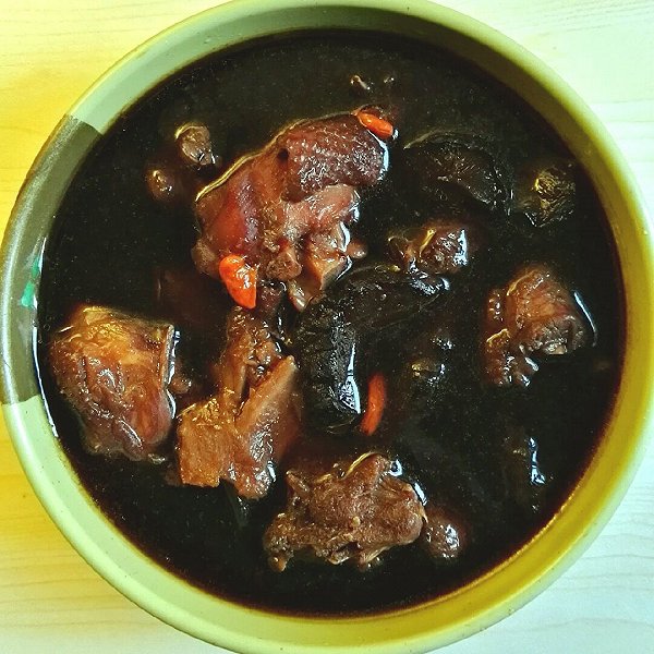 一碗热气腾腾的黑豆煮当归汤,深褐色汤体清澈微浓,表面浮有少量油花,碗中可见煮至裂开的黑豆与浅棕色当归片,旁边配有一双木筷和一小碟红糖,背景为暖色厨房布景,体现家常养生氛围 黑豆煮当归成品汤品展示