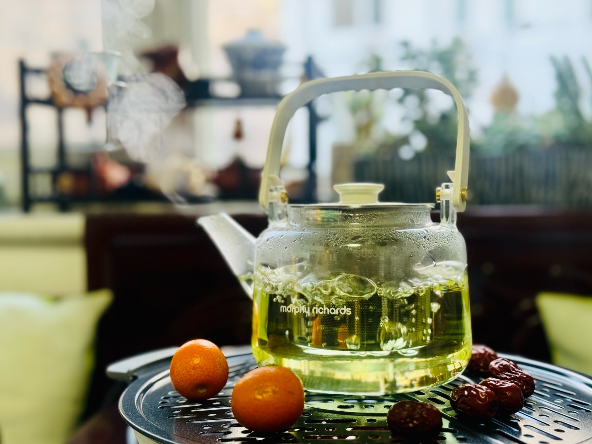 玻璃茶壶中正在冲泡金桔叶茶,壶内漂浮着数片完整的深绿色金桔叶片,水色呈淡黄绿色,散发袅袅热气,旁边摆放干燥的金桔叶样品和陶瓷茶杯,桌面为木质纹理背景 金桔叶茶冲泡过程