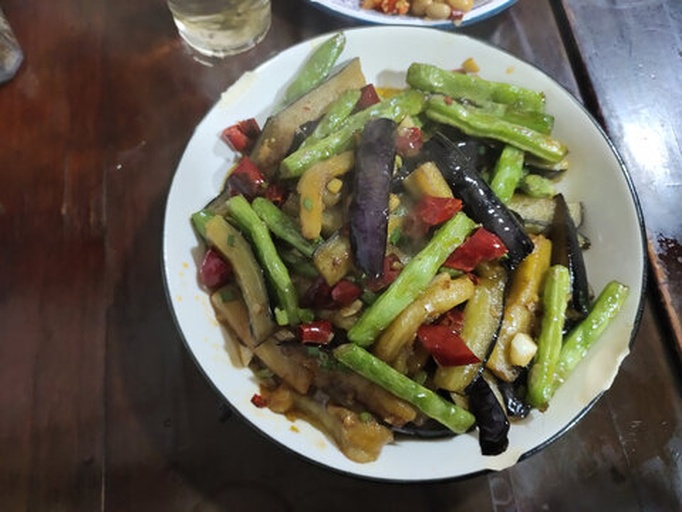 深色炒锅中,茄子条与翠绿毛豆米混合翻炒,汤汁正逐渐收浓,表面泛着油亮光泽,旁边摆放着酱油瓶、蒜末碟等调味容器,背景为家庭厨房环境 锅中正在收汁的毛豆米烧茄子