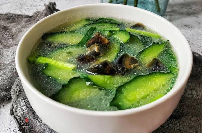 一碗热腾腾的皮蛋冬瓜汤摆放在木质餐桌上,汤色清澈微黄,可见冬瓜片透明柔嫩,点缀着褐色皮蛋块和绿色葱花,旁边配有瓷勺,背景虚化体现家庭厨房氛围 皮蛋冬瓜汤成品展示